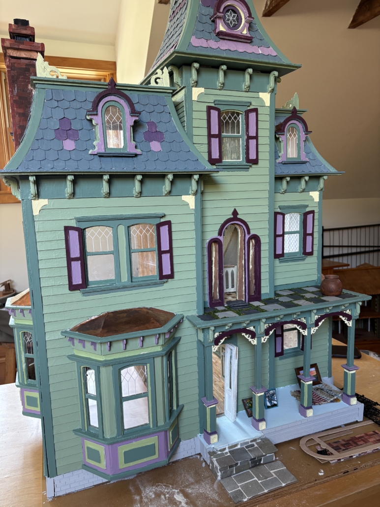 Magnificent Dollhouse created by over 100 local artists. Facade of a Victorian-style dollhouse that is green with bluish roof tiles and purple accents.