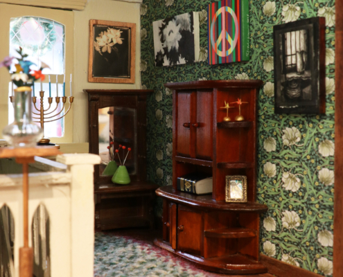 Miniature hallway on the third floor with a mirror desk, a dresser, a menorah, glass vase with flowers, paintings, photographs, paper cranes, and a handwoven rug.