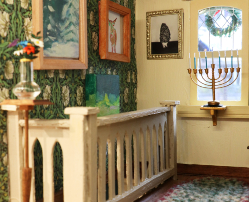 Miniature hallway on the third floor with a mirror desk, a dresser, a menorah, glass vase with flowers, paintings, photographs, paper cranes, and a handwoven rug.