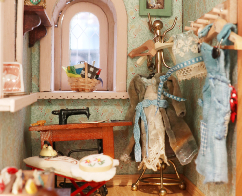 Miniature sewing room with iron and ironing board, thread, paintings, dress, overalls, basket and sewing machine.